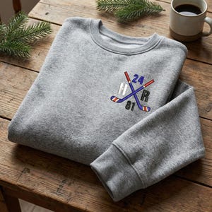 May include: A heather gray sweatshirt with a crew neck and ribbed cuffs. The front features an embroidered hockey-themed design with crossed hockey sticks, the numbers 24 and 81, and the letters H and R in a sporty font. A mug of coffee sits in the background.
