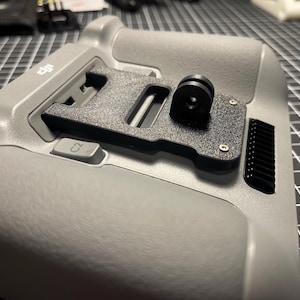 May include: A gray handheld device with a black mounting plate and a black camera mount. The device has a textured surface and a button labeled "C2". The background is a black grid.