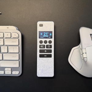 May include: A white remote control with a digital display showing 21 degrees Celsius, buttons, and a keyboard and mouse. The remote is in the center, with the keyboard to the left and the mouse to the right. The background is black.