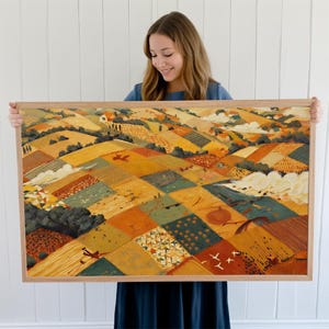 May include: A framed landscape painting with a warm color palette. The artwork depicts a patchwork of fields in shades of yellow, orange, and green, with small houses and trees. The frame is made of light-colored wood.