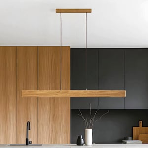 Op de afbeelding: Een rechthoekige houten hanglamp, opgehangen aan het plafond met twee donkere snoeren. De lamp is gemaakt van lichtgekleurd hout en hangt boven een aanrecht. De achtergrond bestaat uit houten kasten en een zwart aanrechtblad.