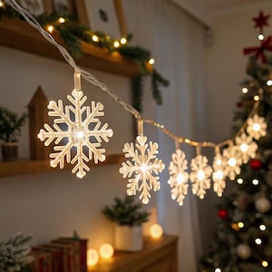 Puede incluir: Una cadena de luces con forma de copo de nieve, iluminadas con un brillo cálido, cuelga sobre un fondo borroso. Las luces están ensartadas en un fino cable plateado, creando un ambiente festivo y decorativo. Los copos de nieve son de un material transparente y texturizado.