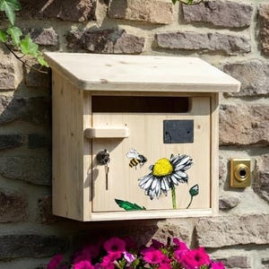 Op de afbeelding: Een houten brievenbus met een schuin dak is bevestigd aan een stenen muur. De brievenbus heeft een geschilderd ontwerp van een bij en madeliefje. Een sleutel hangt aan het slot. Groene klimop en roze bloemen zijn zichtbaar.