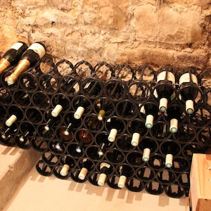 Pode incluir: Um suporte de vinho de metal preto cheio de garrafas de vinho, algumas com folha de ouro. O suporte está embutido em uma parede de pedra, com uma caixa de madeira no chão. O suporte de vinho foi projetado para conter várias garrafas.