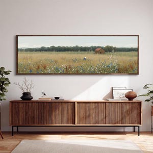 May include: A framed landscape painting depicts a field of wildflowers with a small red-roofed building in the distance. The artwork is encased in a dark brown wooden frame and hangs above a wooden cabinet with a ribbed design.