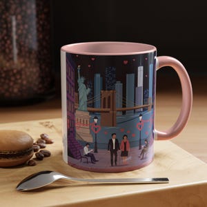 May include: A ceramic mug with a pink handle and interior. The mug features a colorful illustration of the New York City skyline, including the Statue of Liberty, Brooklyn Bridge, and skyscrapers. The design includes couples and hearts.