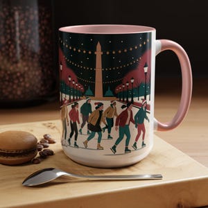 May include: White ceramic mug with a pink interior and handle. The mug features an illustration of people ice skating at night, with a monument and string lights in the background. A macaron and spoon are on the wooden surface.