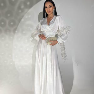 以下が含まれることがあります： 白いサテンのジャンプスーツ。ラップスタイルのボディスとワイドレッグパンツが特徴。長袖は袖口に羽のトリム付き。お揃いのベルトがウエストを締め、ビーズのクラッチバッグが手に持たれています。エレガントでフォーマルな装いです。