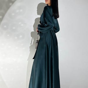 Peut inclure: Longue robe vert fonc&eacute; avec des manches longues et une ceinture assortie. La robe a une jupe fluide et un corsage crois&eacute;. Le mod&egrave;le porte un petit sac &agrave; cha&icirc;ne argent&eacute;. Le fond est un mur blanc uni.