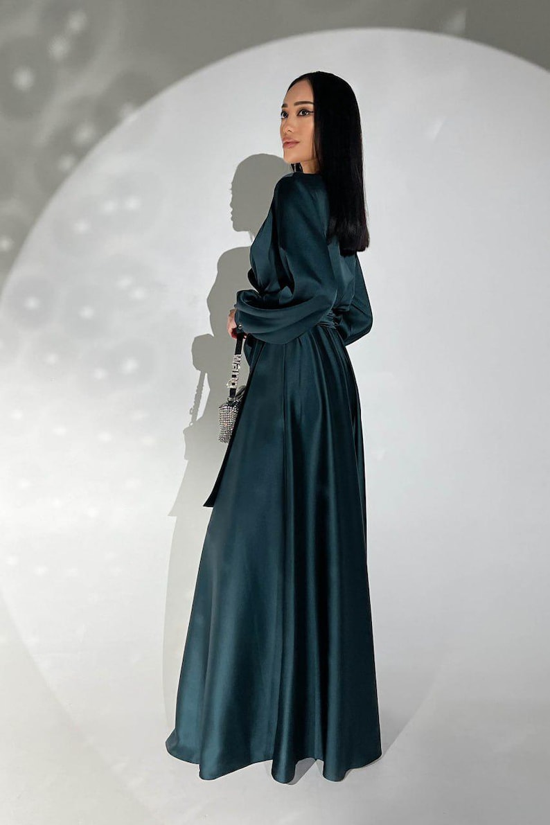 Peut inclure: Longue robe vert fonc&eacute; avec des manches longues et une ceinture assortie. La robe a une jupe fluide et un corsage crois&eacute;. Le mod&egrave;le porte un petit sac &agrave; cha&icirc;ne argent&eacute;. Le fond est un mur blanc uni.