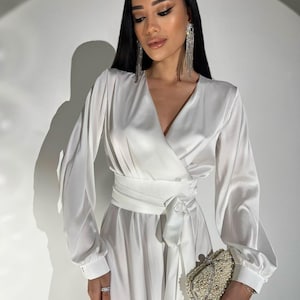 以下が含まれることがあります： 白いサテンのラップドレス、長袖とマッチするベルト付き。 ドレスはビーズのクラッチと組み合わせています。 モデルは長く垂れ下がるイヤリングを着用しています。
