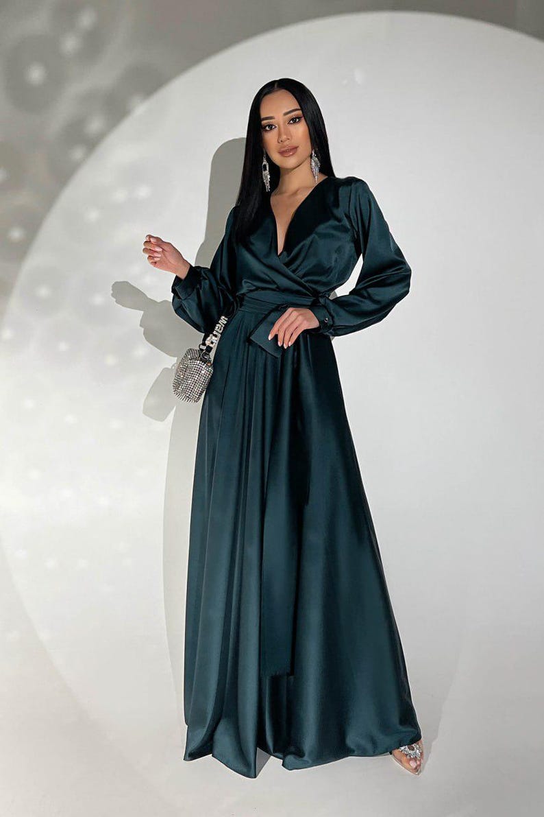 Peut inclure: Longue robe en satin vert fonc&eacute; avec un corsage crois&eacute; et des manches longues. La robe est dot&eacute;e d'une ceinture assortie et d'une jupe fluide. Un petit sac &agrave; main en perles argent&eacute;es est port&eacute; &agrave; l'&eacute;paule. La mod&egrave;le porte des boucles d'oreilles argent&eacute;es.