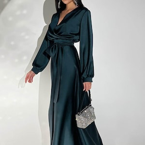 Peut inclure: Une robe portefeuille en satin vert fonc&eacute; avec des manches longues et une ceinture assortie. La robe a un d&eacute;collet&eacute; en V et une jupe longue. Un petit sac &agrave; main rectangulaire argent&eacute; et perl&eacute; est tenu &agrave; la main.