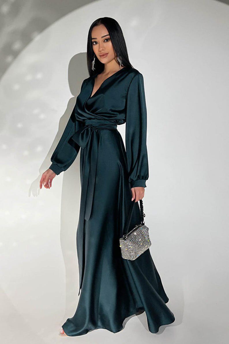 Peut inclure: Une robe portefeuille en satin vert fonc&eacute; avec des manches longues et une ceinture assortie. La robe a un d&eacute;collet&eacute; en V et une jupe longue. Un petit sac &agrave; main rectangulaire argent&eacute; et perl&eacute; est tenu &agrave; la main.