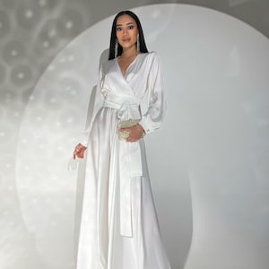 以下が含まれることがあります： ラップスタイルのボディスと長袖の、長く流れるような白いサテンのドレス。 ドレスは、お揃いのウエストタイが特徴で、小さなゴールドのクラッチと組み合わせています。 モデルは長いシルバーのイヤリングを着用しています。