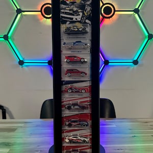Puede incluir: Un expositor negro que muestra una colección de coches de juguete Hot Wheels. El expositor es rectangular con un marco negro y paneles transparentes, que muestran los coches en su embalaje original. El fondo presenta una colorida pantalla de luz LED geométrica.