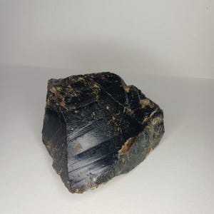 May include: A raw, dark black mineral specimen with a glossy, reflective surface and visible natural striations. The stone features irregular, jagged edges and subtle brownish-tan inclusions embedded within its dense, opaque structure.