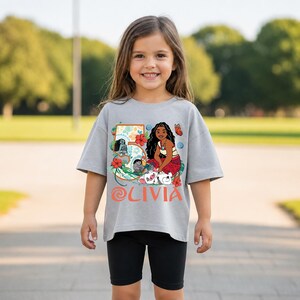 May include: A gray t-shirt featuring a colorful graphic of a cartoon character, flowers, and the number 5. The name "OLIVIA" is printed in coral-colored letters below the graphic. The shirt is worn by a young girl.