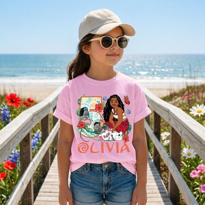 May include: A pink t-shirt featuring a colorful graphic of a cartoon character, flowers, and the name "Olivia". The shirt is worn by a person wearing sunglasses and a beige cap, standing on a wooden walkway near the beach.