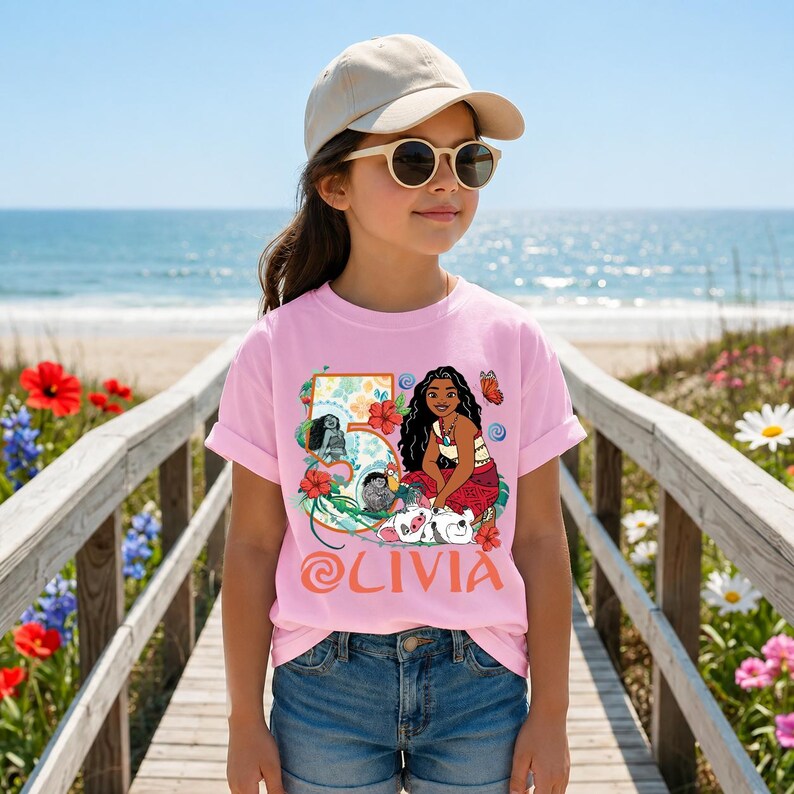 May include: A pink t-shirt featuring a colorful graphic of a cartoon character, flowers, and the name "Olivia". The shirt is worn by a person wearing sunglasses and a beige cap, standing on a wooden walkway near the beach.