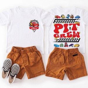 Puede incluir: Se muestran dos camisetas blancas con gr&aacute;ficos de coches y pantalones cortos marrones con zapatos sin cordones a cuadros. Una camiseta presenta un coche de carreras rojo con el nombre "Alex". La otra camiseta dice "Pit Crew" en letras rojas.