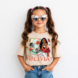 May include: A tan t-shirt featuring a colorful graphic of a cartoon character, flowers, and the name "Olivia." The graphic includes the number "5." The girl in the image is wearing heart-shaped sunglasses and blue jeans.