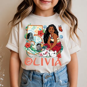 May include: Cream-colored t-shirt featuring a colorful graphic of a cartoon character with long black hair, a red skirt, and a necklace. The number 5 is in a floral design, and the name "OLIVIA" is printed in orange. A butterfly and other characters are also included.