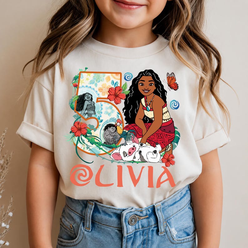 May include: Cream-colored t-shirt featuring a colorful graphic of a cartoon character with long black hair, a red skirt, and a necklace. The number 5 is in a floral design, and the name "OLIVIA" is printed in orange. A butterfly and other characters are also included.