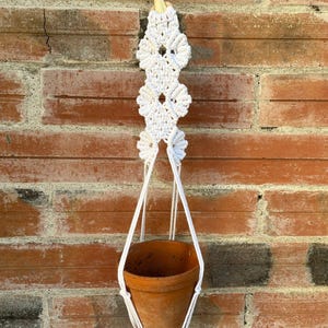 Può includere: Un portavaso in macramè bianco con un vaso in terracotta. Il design in macramè presenta nodi a forma di diamante e un piolo di legno per appendere. Il vaso in terracotta è vuoto, pronto per una pianta.