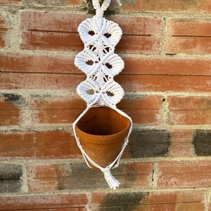Peut inclure: Un porte-plante en macramé blanc avec un pot en terre cuite. Le motif en macramé présente une série de losanges interconnectés. Le pot est suspendu au macramé, accroché à un mur de briques rouges.