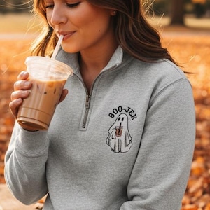 May include: A light gray quarter-zip pullover sweatshirt featuring an embroidered graphic on the left chest of a white ghost holding a brown iced coffee cup with the text BOO-JEE arched above the ghost in black lettering.