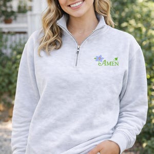 Könnte beinhalten: Hellgraues Sweatshirt mit einem Viertel-Reißverschluss am Ausschnitt. Das Sweatshirt hat eine blaue Blume und das Wort "AMEN" in grüner Schrift. Das Model trägt das Sweatshirt mit einer khakifarbenen Hose.