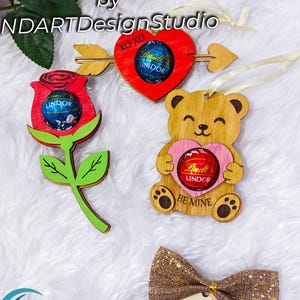 May include: Wooden laser-cut Valentine's Day decorations. Includes a red heart with an arrow, a rose, a teddy bear holding a heart, and a bow. Each piece holds a Lindor chocolate. The image also includes the text "Digital Download by NDART Design Studio."