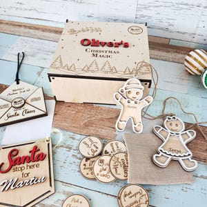 Könnte beinhalten: Ein hölzernes Weihnachts-Box-Set mit dem Text "Oliver's Christmas Magic". Enthält Lebkuchen-Ornamente, ein Santa-Schild und runde Token mit festlichen Phrasen. Das Set befindet sich auf einer hellblau-weißen Holzoberfläche.