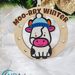 May include: Wooden laser-cut ornament featuring a cartoon cow wearing a blue hat, with the text "MOO-RRY WINTER". The ornament is circular with snowflake accents. The design is a digital download by NDARTDesignStudio.