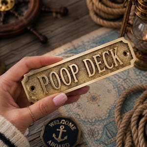 Könnte beinhalten: Ein rechteckiges Messingschild mit einer verwitterten, strukturierten Oberfläche und dem erhabenen, geprägten Text POOP DECK in Großbuchstaben. Das Schild hat abgerundete Ecken und zwei vorgebohrte Schraublöcher an der linken und rechten Seite.