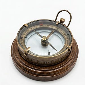 Puede incluir: Una brújula redonda de latón con cubierta de cristal, que presenta una esfera blanca con puntos cardinales y marcas de grados en negro. La brújula está montada sobre una base circular de madera marrón oscura con el texto grabado OUR JOURNEY BEG.