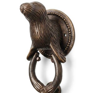 May include: A bronze-toned metal door knocker featuring a sculpted sea otter figure attached to a circular mounting plate. The otter's front flippers hold a curved, ring-shaped knocker that hangs downward, showcasing a textured, weathered finish.