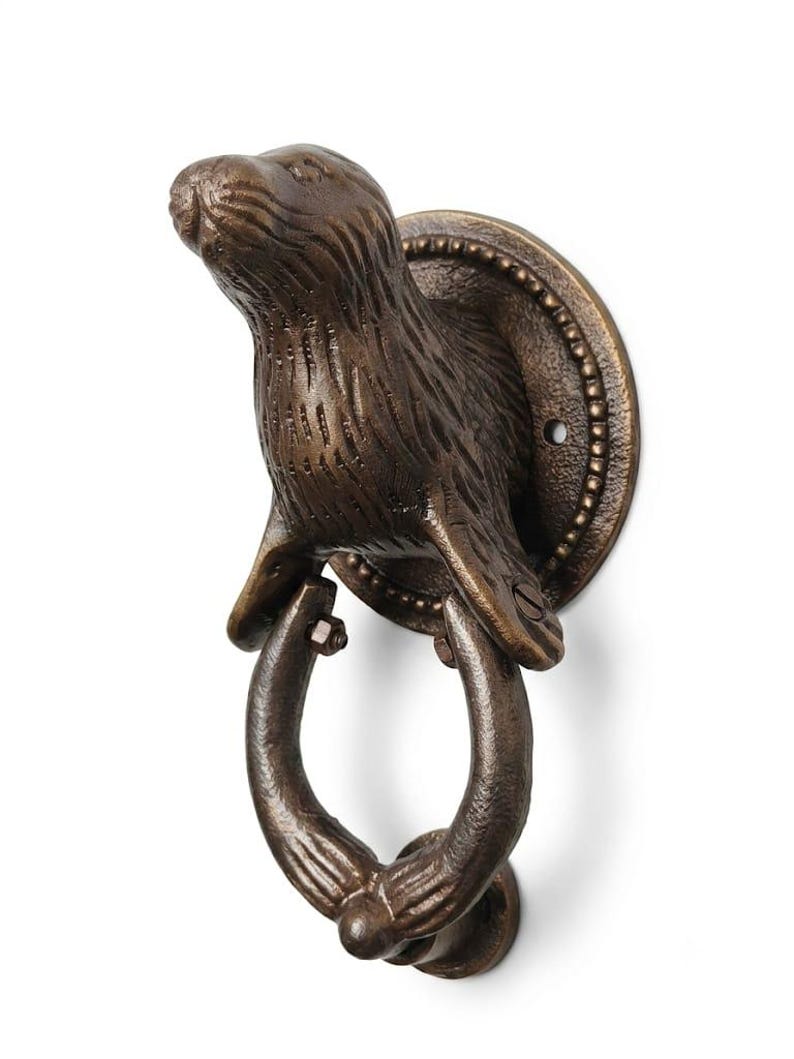 May include: A bronze-toned metal door knocker featuring a sculpted sea otter figure attached to a circular mounting plate. The otter's front flippers hold a curved, ring-shaped knocker that hangs downward, showcasing a textured, weathered finish.