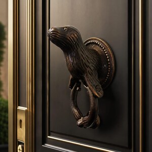 May include: A bronze-toned metal door knocker shaped like an otter, featuring a textured fur finish and a circular ring held by the otter's front paws. The knocker is mounted on a circular base plate and attached to a matte black door with gold trim.