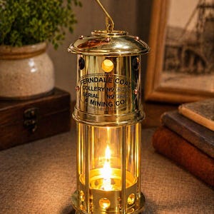 Op de afbeelding: Een messing mijnwerkerslamp met een brandende vlam erin. De lantaarn heeft een haak om op te hangen en een glazen behuizing. De tekst "Ferndale Coal Colliery" is zichtbaar op de zijkant. De lantaarn is ongeveer 30 cm hoog.