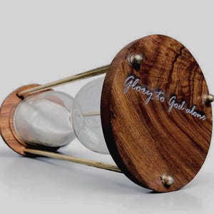 May include: An hourglass with wooden end pieces and a clear glass center. The hourglass is filled with white sand. The text "Glory to God alone" is engraved on the wooden end piece. The hourglass is a decorative item.
