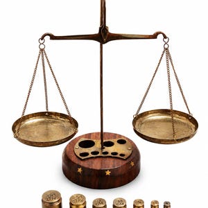 May include: A brass balance scale with two hanging pans suspended by chains from a central beam. The scale features a circular wooden base with inlaid star accents and a brass weight holder. Seven cylindrical brass weights are arranged in front.