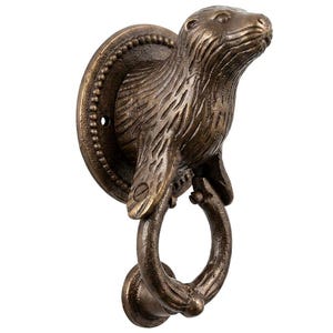 Puede incluir: Un llamador de puerta de metal en tono bronce con la cabeza y el torso de una nutria esculpidos, unidos a una placa de montaje circular con borde de cuentas. Un anillo circular texturizado cuelga de las patas delanteras de la nutria.