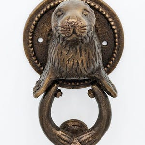 May include: A bronze-toned metal door knocker featuring a sculpted seal head mounted on a circular backplate with a beaded edge. The seal's front flippers extend downward to hold a U-shaped ring, which serves as the swinging part of the knocker.
