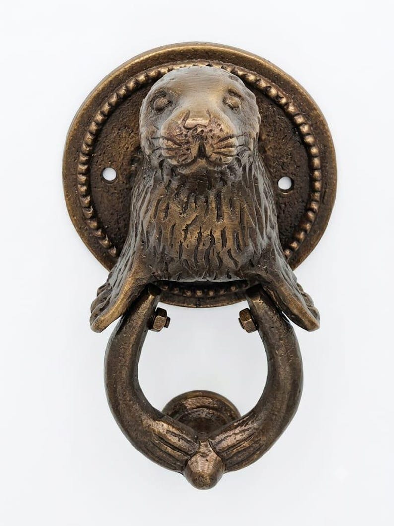 May include: A bronze-toned metal door knocker featuring a sculpted seal head mounted on a circular backplate with a beaded edge. The seal's front flippers extend downward to hold a U-shaped ring, which serves as the swinging part of the knocker.