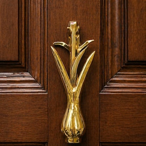 Puede incluir: Un llamador de puerta dorado con un diseño floral estilizado, montado en una puerta de madera marrón oscuro. El llamador tiene una base bulbosa y elementos alargados en forma de hoja.