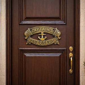 Pode incluir: Uma porta de madeira castanho-escura com uma placa de metal cor de latão com o texto CAPTAINS QUARTERS. A placa é oval, decorada com cabeças de peixe estilizadas nas laterais e um ícone de âncora central, montada no centro do painel.