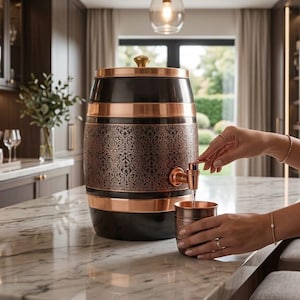 Puede incluir: Un dispensador de bebidas con forma de barril en negro y cobre con una banda central que presenta un intrincado patrón floral. El dispensador tiene un grifo de color cobre y una tapa a juego con un pequeño pomo, junto a una taza a juego.
