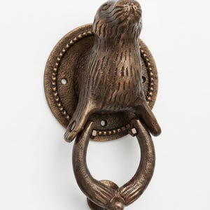 May include: A bronze-colored metal door knocker featuring a sculpted seal head mounted on a circular backplate with a beaded edge. The seal's front flippers extend downward to hold a circular ring that serves as the striking part of the mechanism.